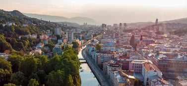 An elevated view of Sarajevo in Bosnia and Herzegovina with the Miljacka river flowing between urban buildings and green hillsides under a warm sunset, representing the established range of business entities in the region.